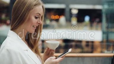 迷人的年轻女子一手拿着智能手机，一手拿着咖啡坐在咖啡馆里